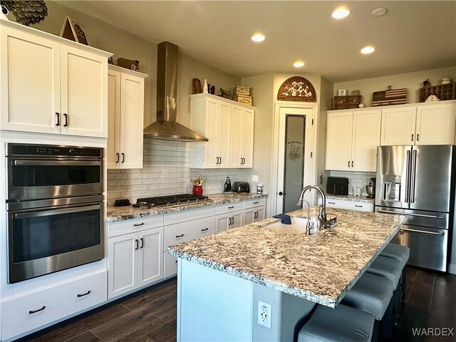 kitchen featuring stainless steel appliances, a sink, white cabinetry, wall chimney range hood, and dark wood-style floors