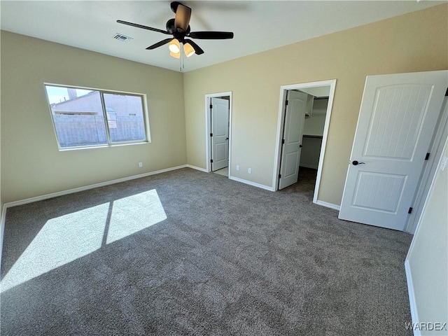 unfurnished bedroom featuring a walk in closet, visible vents, dark carpet, a ceiling fan, and baseboards