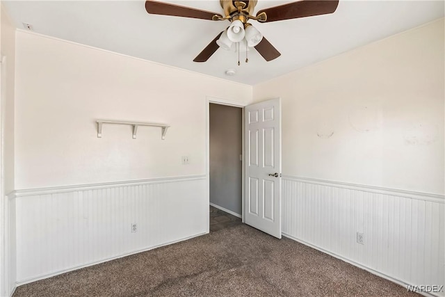 spare room with ceiling fan, dark colored carpet, and wainscoting