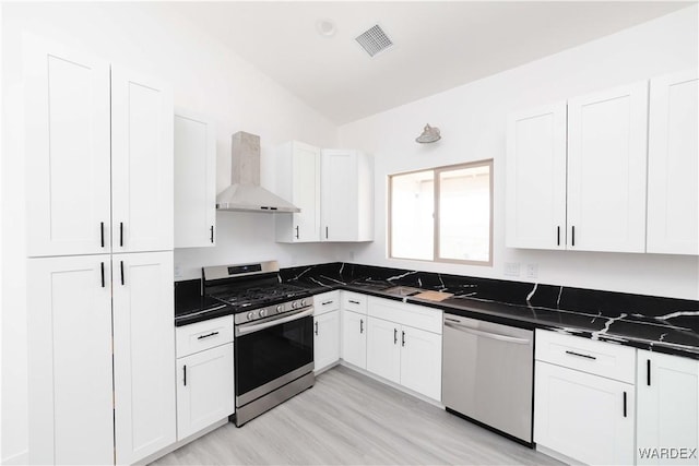 kitchen featuring stainless steel appliances, visible vents, white cabinets, wall chimney range hood, and dark stone countertops