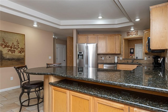 kitchen with a tray ceiling, stainless steel appliances, backsplash, a sink, and dark stone countertops