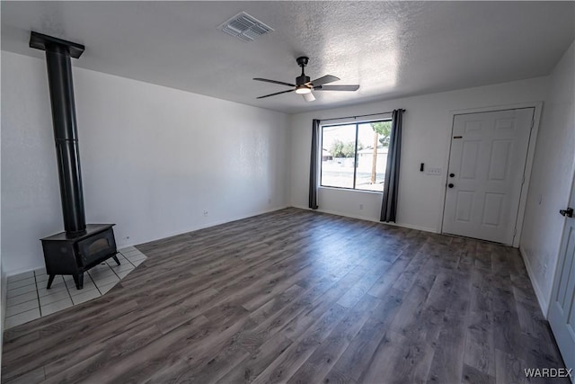 unfurnished living room featuring visible vents, ceiling fan, dark wood-type flooring, a wood stove, and a textured ceiling