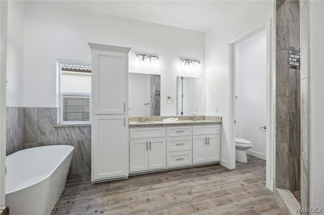 bathroom with double vanity, toilet, a freestanding bath, wood finish floors, and tile walls