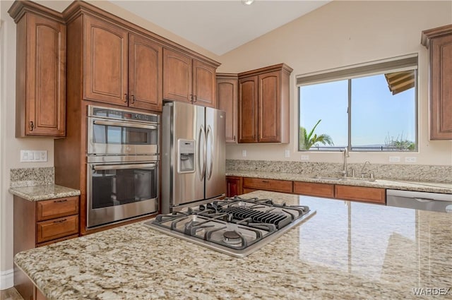 kitchen featuring light stone counters, brown cabinets, appliances with stainless steel finishes, vaulted ceiling, and a sink