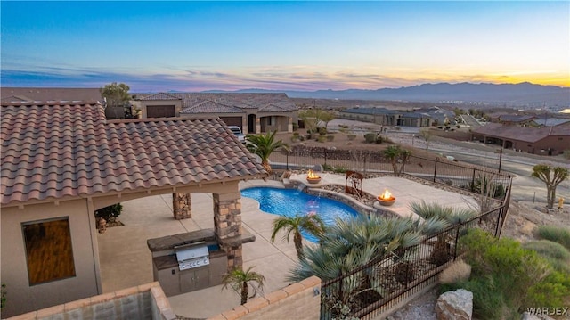 pool at dusk featuring an outdoor kitchen, a fenced in pool, a patio, a fenced backyard, and a mountain view