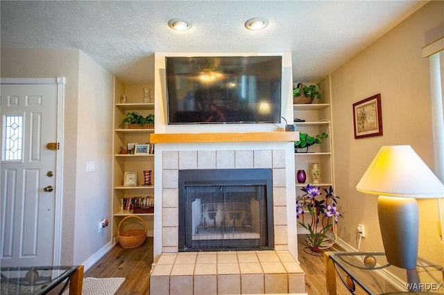 living room with a textured ceiling, built in shelves, a tiled fireplace, and wood finished floors