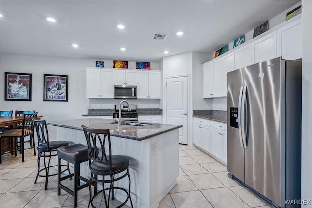 kitchen featuring stainless steel appliances, visible vents, white cabinets, dark stone counters, and a center island with sink