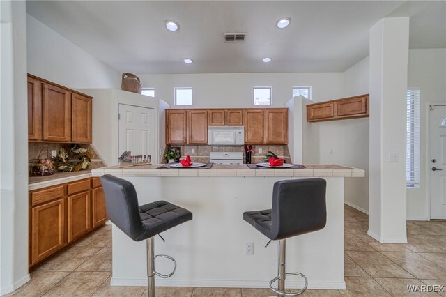 kitchen featuring brown cabinetry, tile countertops, white microwave, a breakfast bar area, and stove