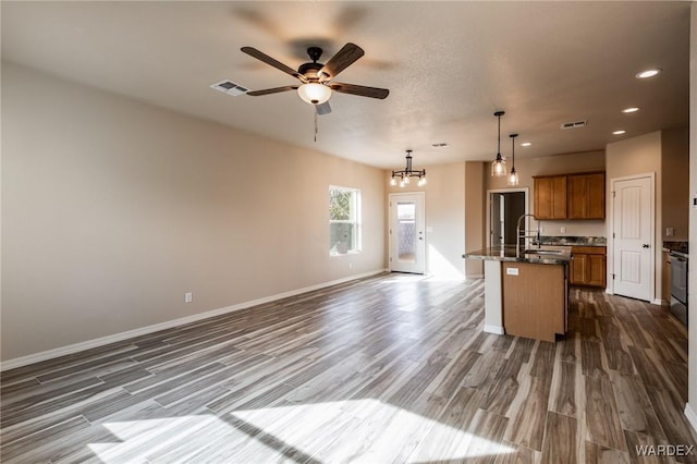 kitchen with a center island with sink, dark countertops, visible vents, brown cabinetry, and a sink