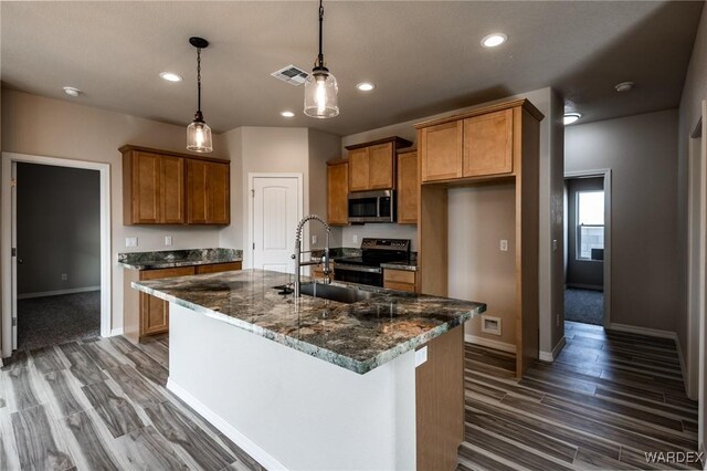 kitchen featuring brown cabinets, stainless steel microwave, hanging light fixtures, a kitchen island with sink, and a sink