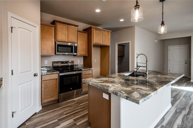 kitchen with stainless steel appliances, a sink, brown cabinetry, an island with sink, and decorative light fixtures