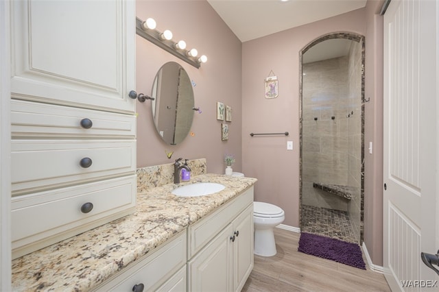 full bathroom with toilet, a tile shower, vanity, wood finished floors, and baseboards