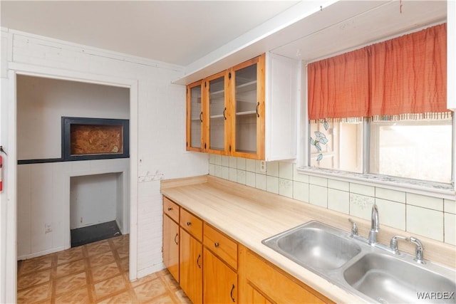 kitchen with brown cabinetry, glass insert cabinets, a sink, light countertops, and backsplash