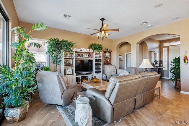 living area featuring a ceiling fan, arched walkways, visible vents, and a lit fireplace