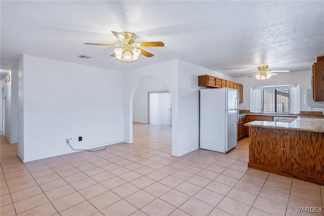 kitchen featuring arched walkways, a ceiling fan, brown cabinets, freestanding refrigerator, and a peninsula