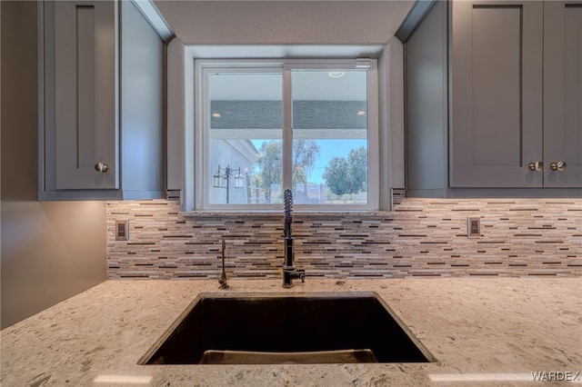 kitchen with a sink, light stone counters, backsplash, and gray cabinetry