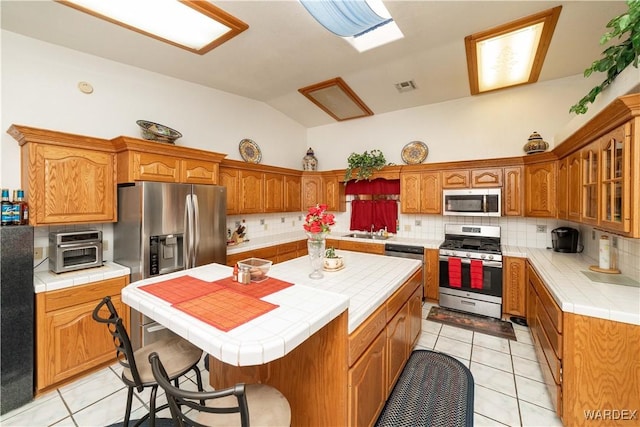 kitchen featuring tile counters, a kitchen island, appliances with stainless steel finishes, brown cabinets, and a sink