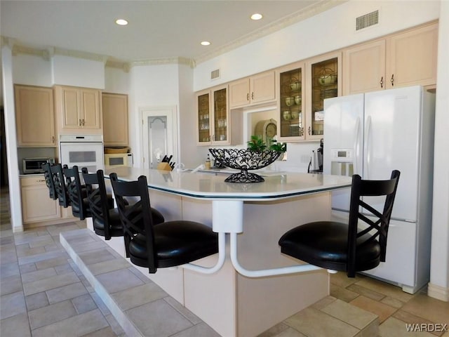 kitchen with light countertops, light brown cabinetry, glass insert cabinets, white appliances, and a kitchen bar