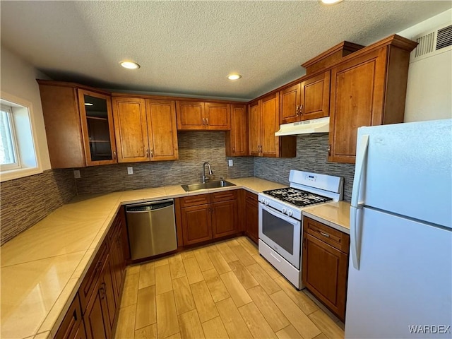 kitchen featuring under cabinet range hood, white appliances, a sink, light countertops, and brown cabinetry