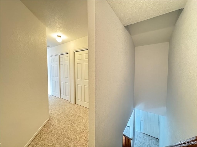 hall with light colored carpet, a textured wall, a textured ceiling, and baseboards