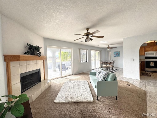 living room with arched walkways, a tiled fireplace, light tile patterned flooring, ceiling fan, and a textured ceiling