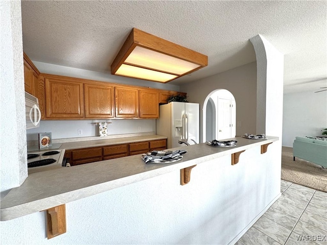 kitchen featuring a breakfast bar area, light countertops, brown cabinetry, white appliances, and a peninsula