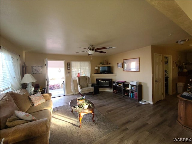 living room featuring dark wood-type flooring, a glass covered fireplace, a ceiling fan, and baseboards