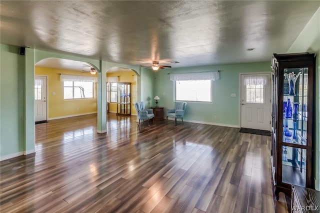 unfurnished living room with arched walkways, ceiling fan, dark wood-style floors, and baseboards