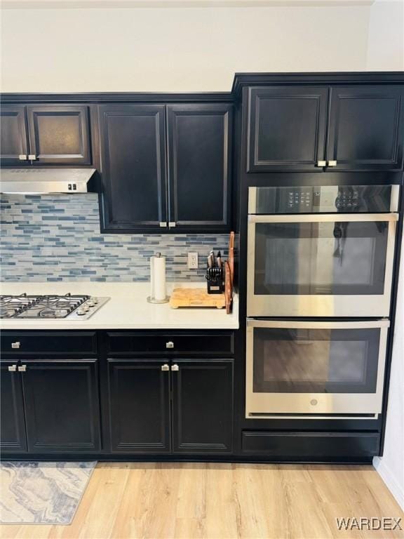 kitchen with appliances with stainless steel finishes, light wood finished floors, light countertops, tasteful backsplash, and under cabinet range hood