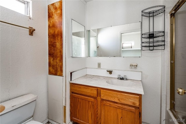 bathroom featuring a textured wall, shower / bath combination with glass door, vanity, and toilet