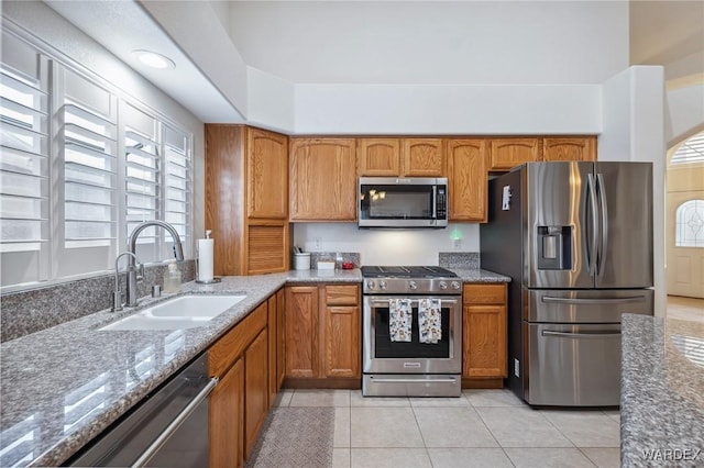 kitchen with light tile patterned floors, brown cabinetry, appliances with stainless steel finishes, light stone countertops, and a sink