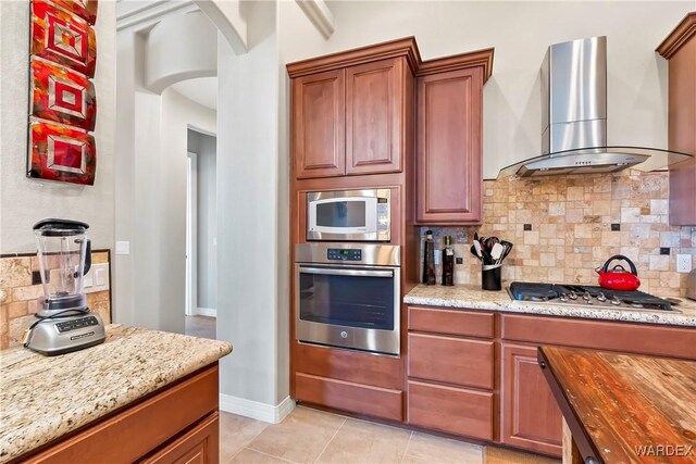 kitchen featuring light stone counters, arched walkways, stainless steel appliances, backsplash, and wall chimney range hood