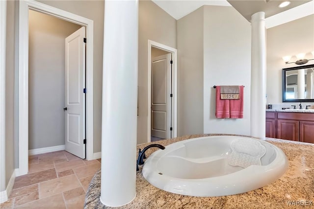 bathroom with stone tile floors, decorative columns, baseboards, a garden tub, and vanity