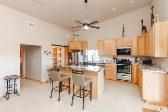 kitchen with a barn door, a kitchen island, appliances with stainless steel finishes, light stone countertops, and light brown cabinetry