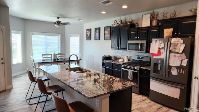 kitchen featuring visible vents, an island with sink, dark cabinets, stainless steel appliances, and a sink