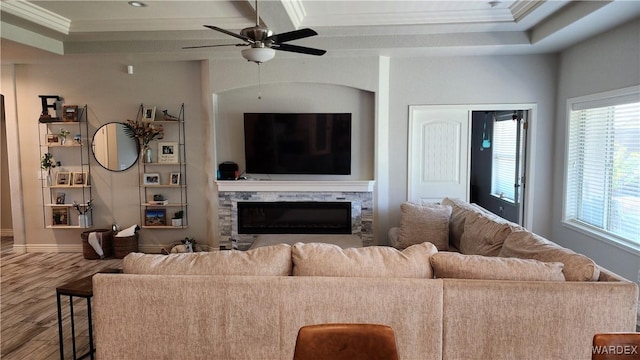 living area with ceiling fan, a stone fireplace, wood finished floors, ornamental molding, and a raised ceiling