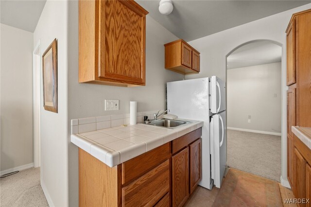 kitchen with tile countertops, arched walkways, light carpet, a sink, and brown cabinetry