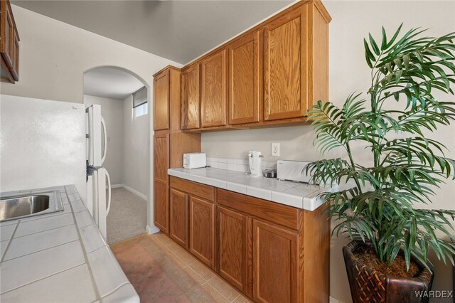 kitchen with arched walkways, brown cabinets, light tile patterned floors, tile counters, and a sink