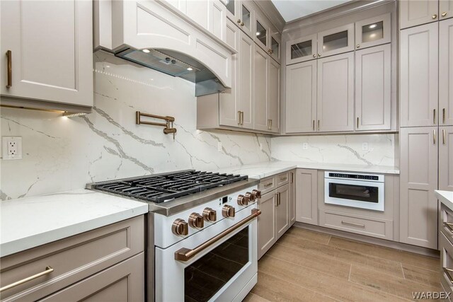 kitchen featuring light stone countertops, tasteful backsplash, gas stove, glass insert cabinets, and custom range hood