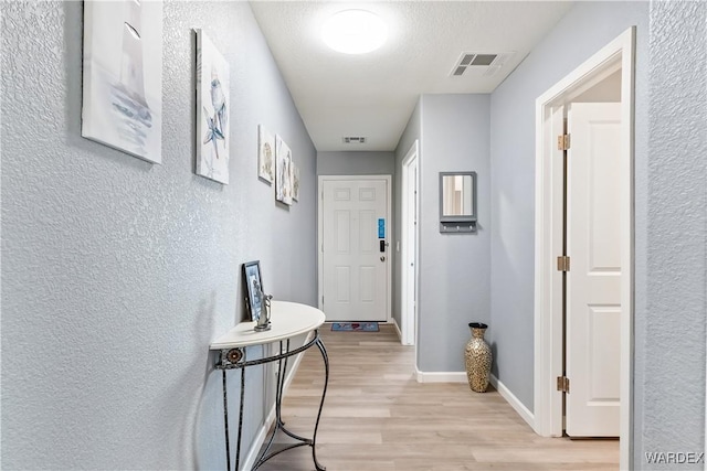 hallway featuring light wood finished floors, baseboards, visible vents, and a textured wall