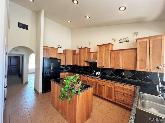 kitchen featuring arched walkways, black refrigerator with ice dispenser, a kitchen island, a sink, and dark stone countertops