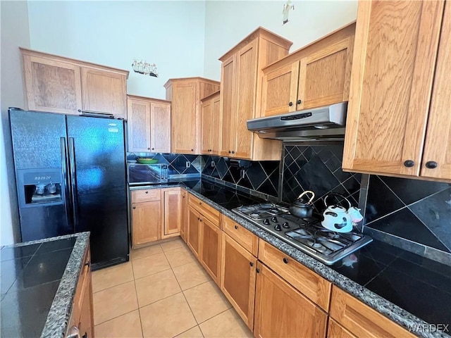 kitchen with light tile patterned floors, under cabinet range hood, stainless steel gas cooktop, black fridge with ice dispenser, and dark stone counters