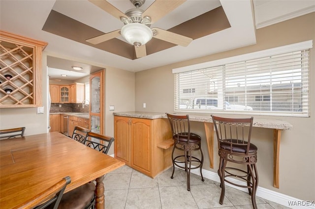 kitchen featuring decorative backsplash, glass insert cabinets, brown cabinetry, a ceiling fan, and light tile patterned flooring