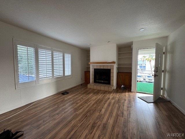 unfurnished living room with built in shelves, dark wood finished floors, a textured ceiling, a tile fireplace, and baseboards