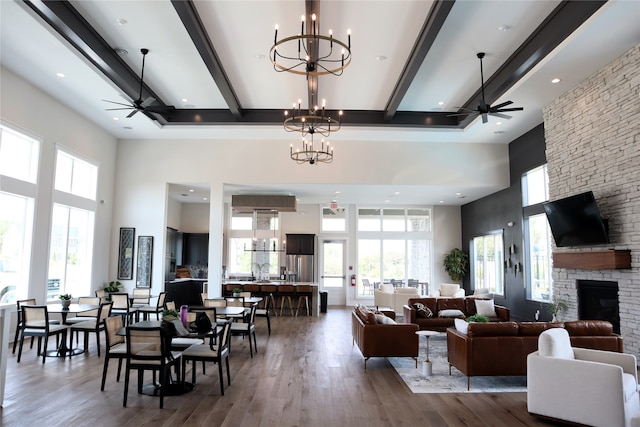 interior space featuring beam ceiling, a stone fireplace, hardwood / wood-style flooring, a high ceiling, and ceiling fan with notable chandelier