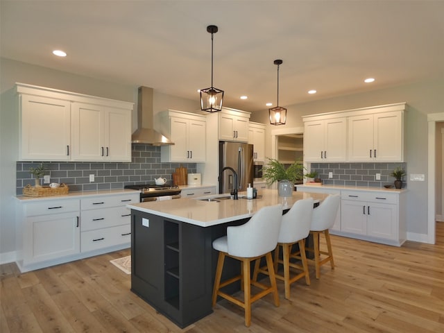 kitchen with white cabinets, light hardwood / wood-style flooring, sink, wall chimney range hood, and a center island with sink