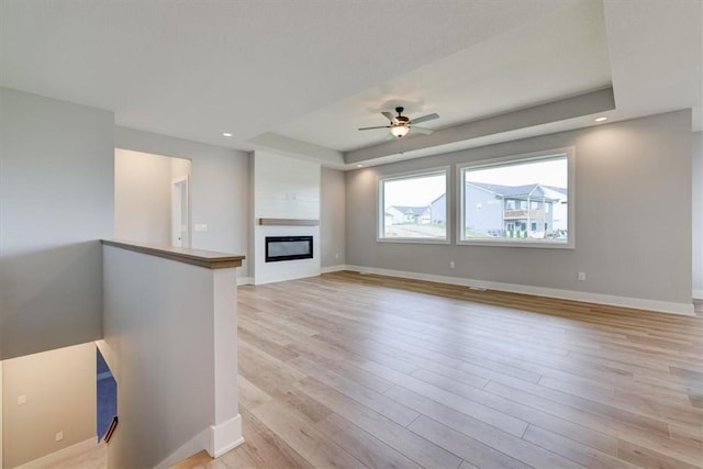 unfurnished living room featuring a raised ceiling, ceiling fan, a fireplace, and light wood-type flooring