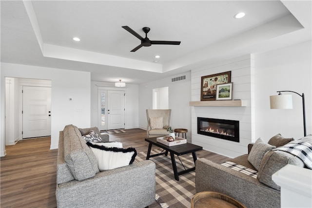 living room featuring ceiling fan, a large fireplace, a tray ceiling, and dark hardwood / wood-style flooring