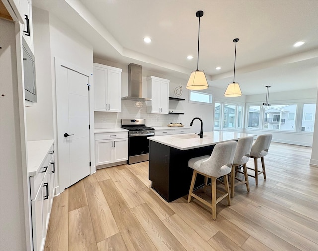 kitchen featuring stainless steel range with gas stovetop, white cabinets, a kitchen island with sink, and wall chimney exhaust hood