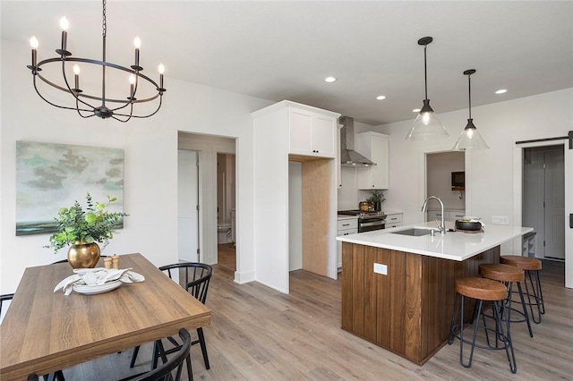 kitchen featuring wall chimney range hood, sink, hanging light fixtures, an island with sink, and white cabinetry